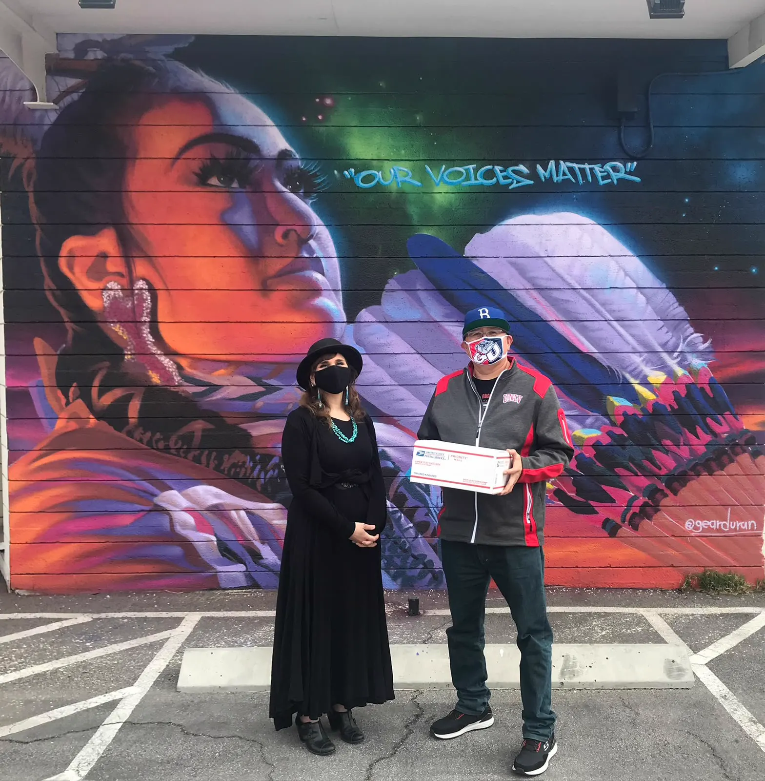 Two people standing before a large mural, one holding a postal box
