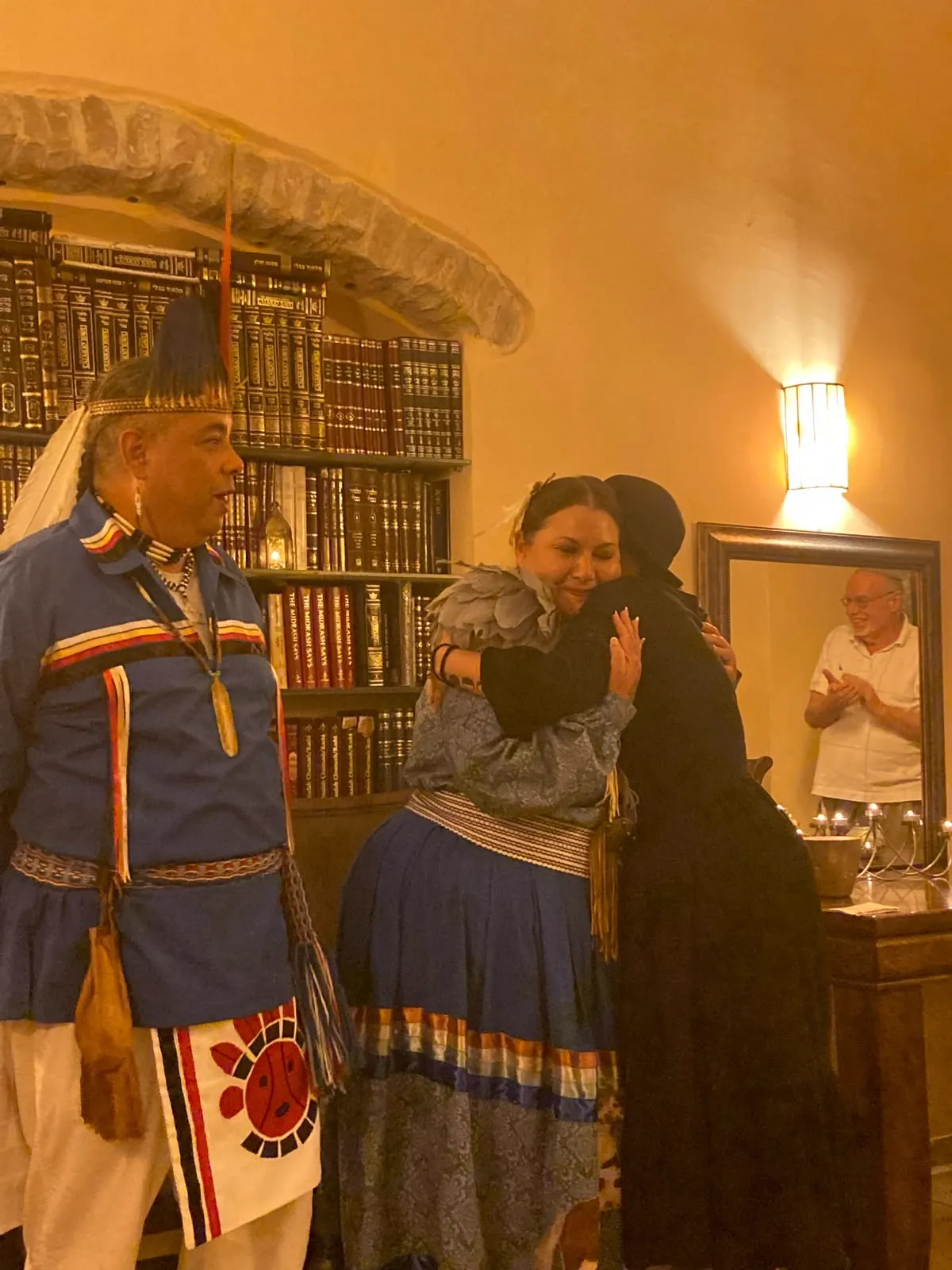 Two people embracing beside a bookshelf while a man in traditional dress stands nearby